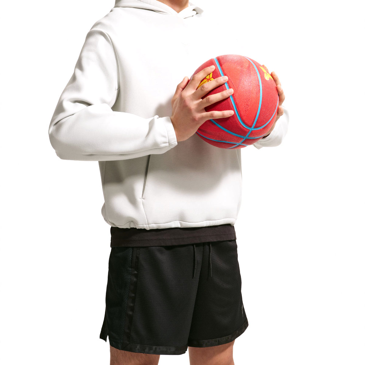 A person wearing a white pullover hoodie and black athletic shorts holds a red basketball with blue and yellow lines against a plain white background.