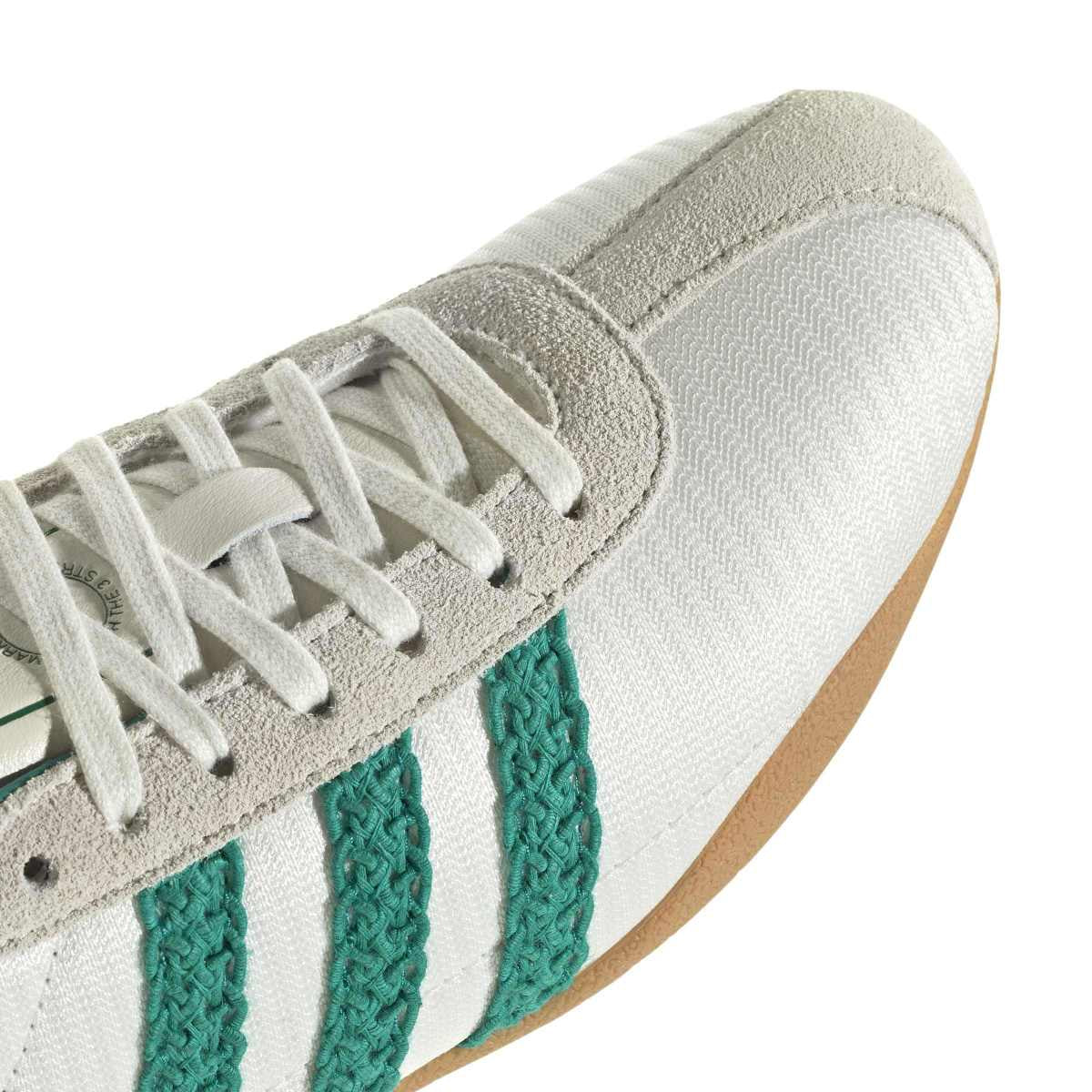 Close-up of a white and light gray sneaker with three green knit stripes and a tan outsole, shown on a white background.