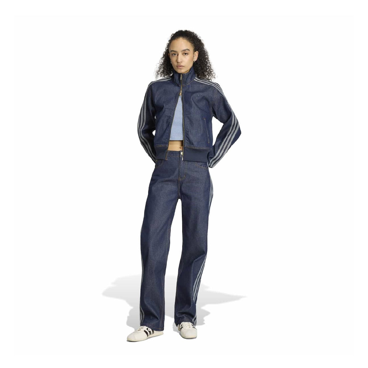 A model stands against a white studio background wearing a dark blue denim tracksuit with white stripes on the sleeves and legs, paired with a light blue crop top and white sneakers.