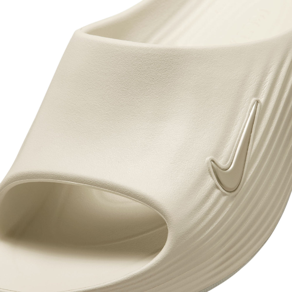 A close-up of a Nike slide sandal in a light cream or off-white color, showing the textured foam upper and embossed Swoosh logo on a white background.