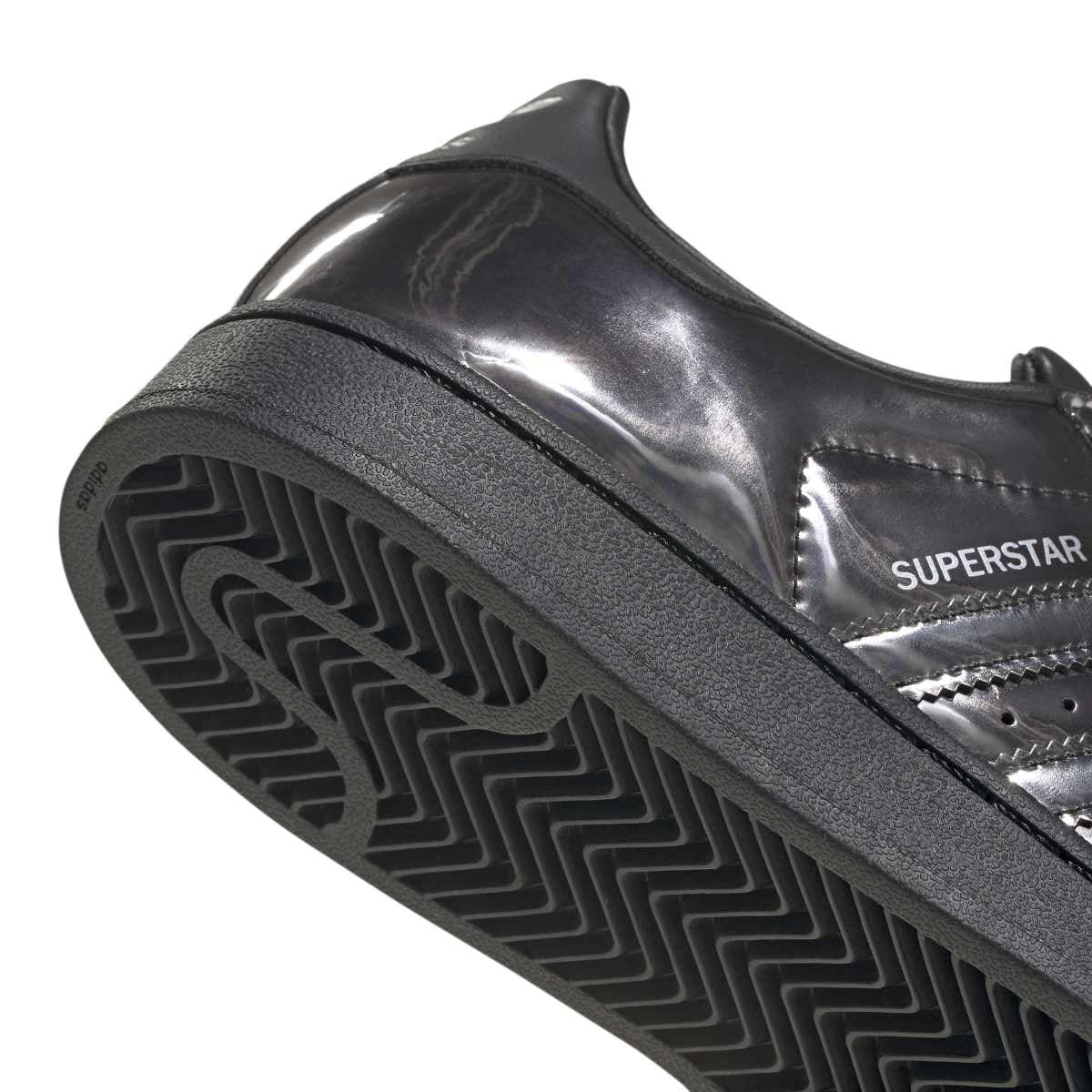 Close-up of a black Adidas Superstar sneaker showing the glossy patent leather side, stitching, and the textured black rubber sole with zigzag tread.