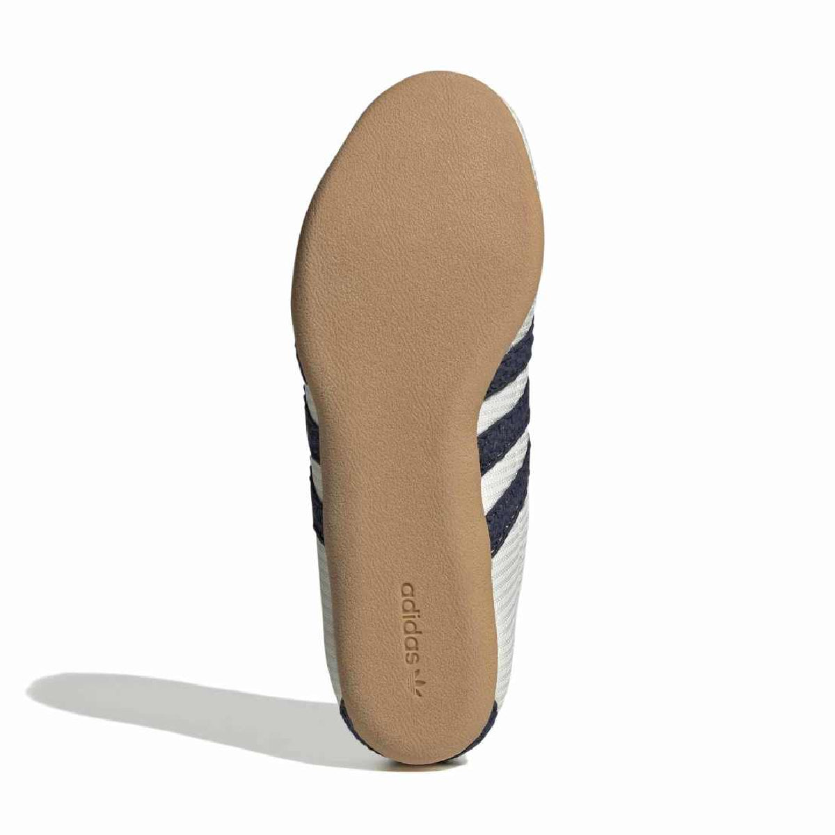 The outsole of an Adidas sneaker, showing a tan rubber sole with the Adidas logo printed on it, alongside white and navy blue stripes on the side, set against a plain white background.