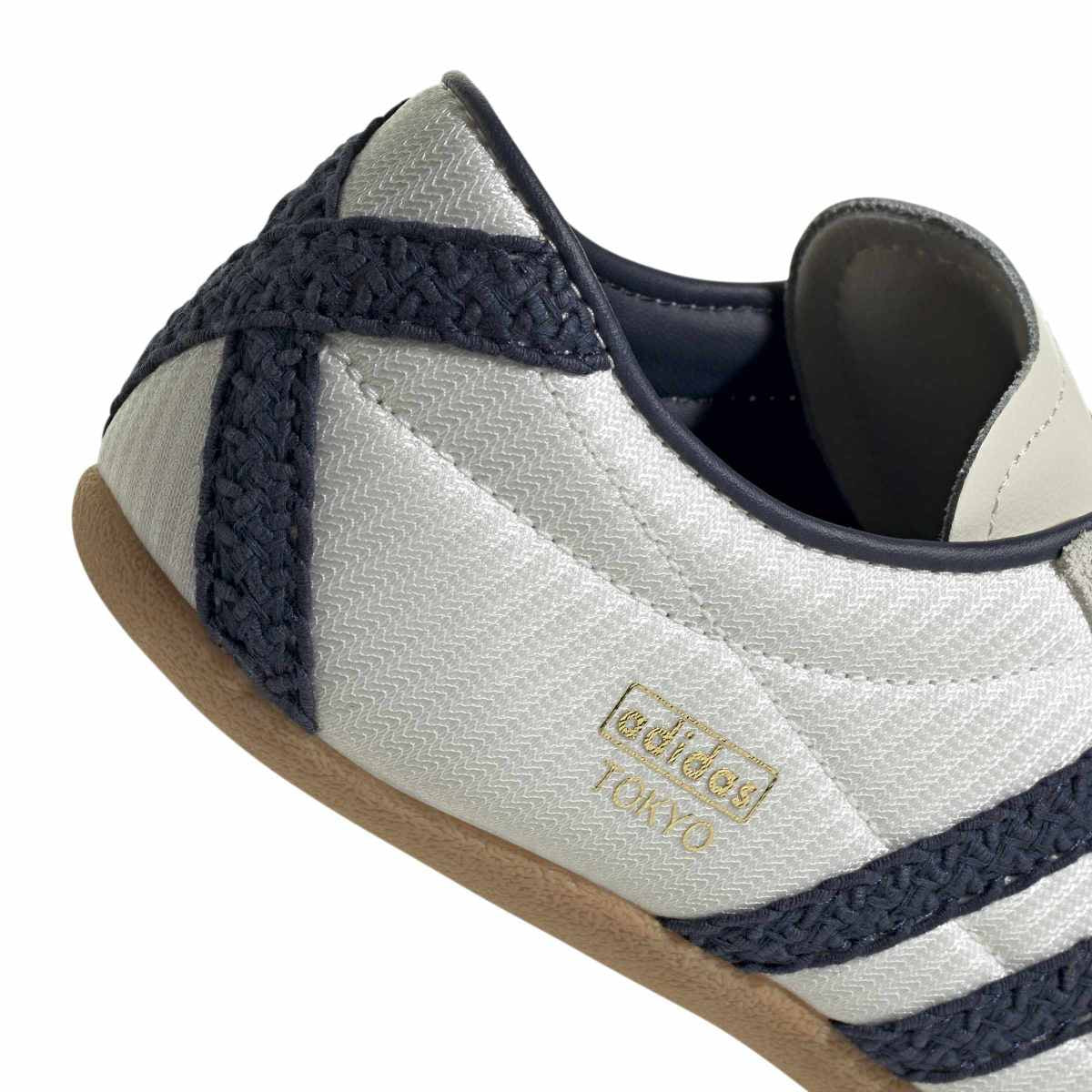 Close-up of an Adidas Tokyo sneaker showing the side panel with textured white fabric, navy blue braided stripes, a tan rubber sole, and gold 'adidas TOKYO' branding.