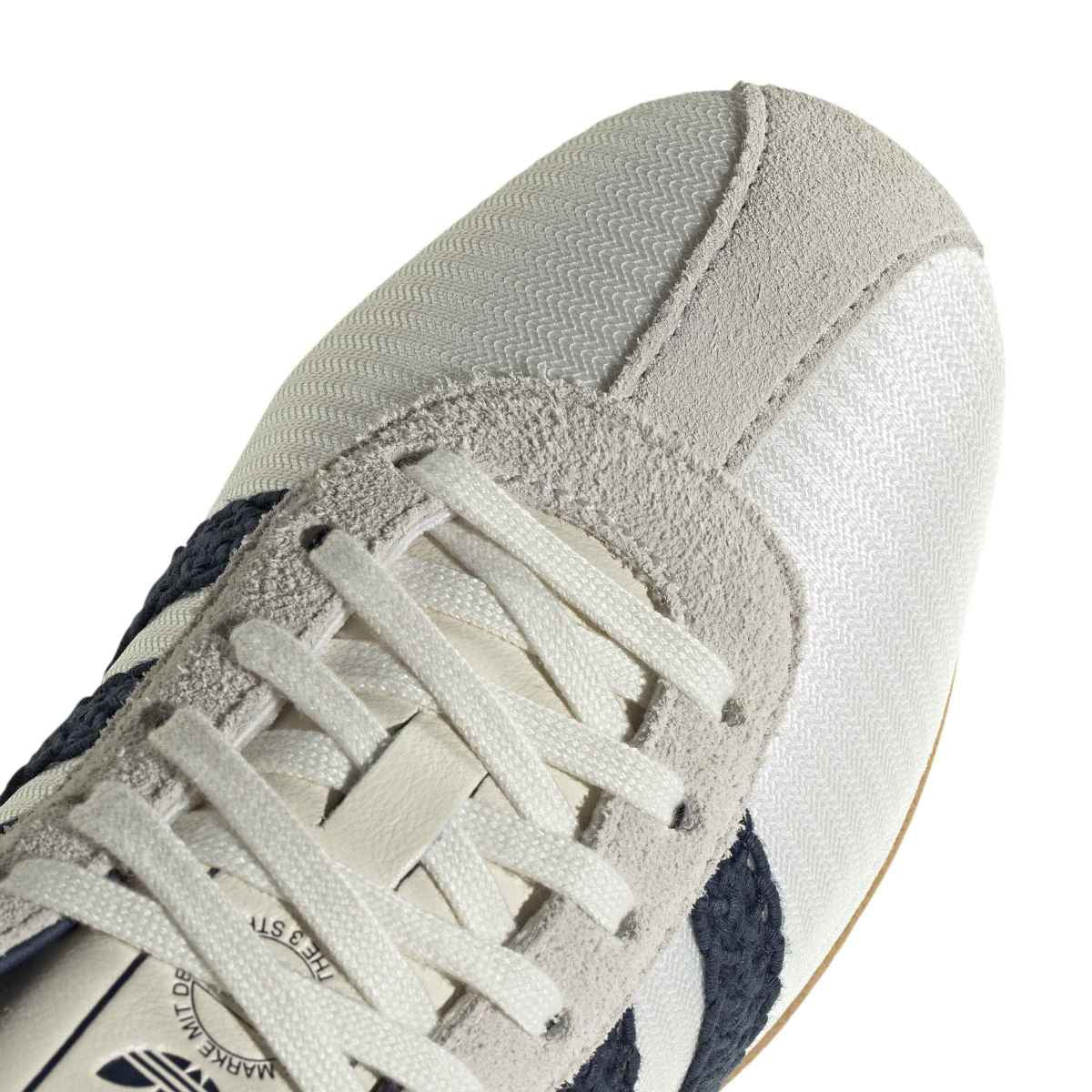Close-up of a low-top sneaker showing the toe box, laces, and textured side panels in off-white, beige, and dark navy.
