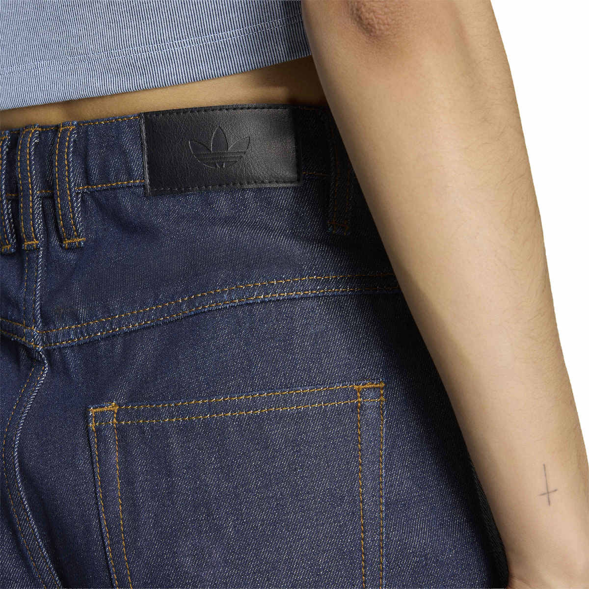 Close-up of a person wearing dark indigo denim jeans with visible waistband stitching, a black leather Adidas logo patch, and contrast gold-toned stitching. The model's arm is visible on the right side against a white background.