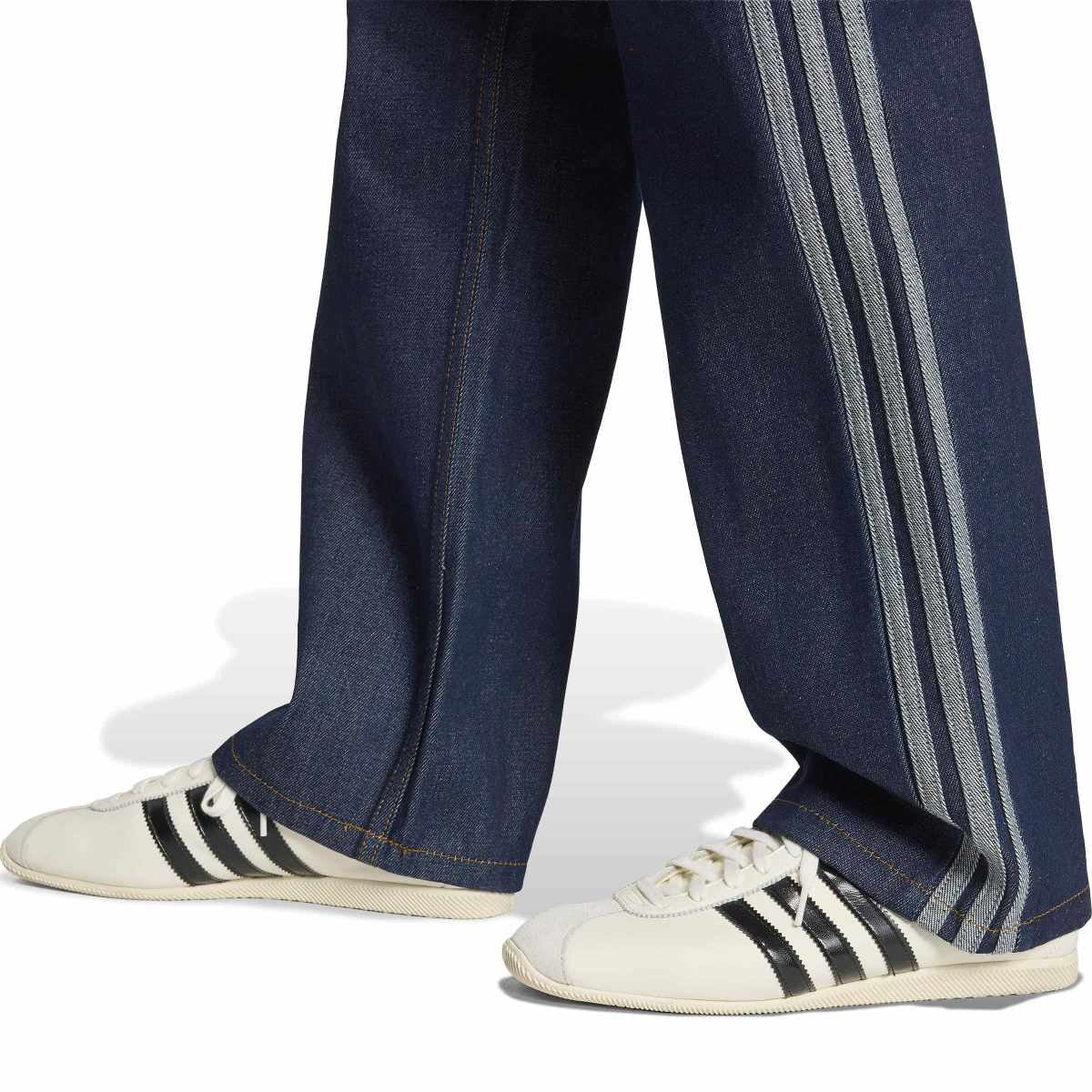 Close-up of a person's legs wearing dark blue denim track pants with three white stripes down the side and white low-top sneakers with black stripes, shown against a white background.